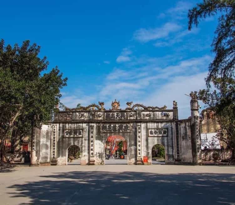 Ancient Vietnamese temple gate with traditional architecture, lush trees, and clear blue sky, representing cultural heritage.